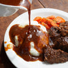 Plated meal with mashed potatoes, beef, carrots, and gravy being poured over the dish on a wooden table.
