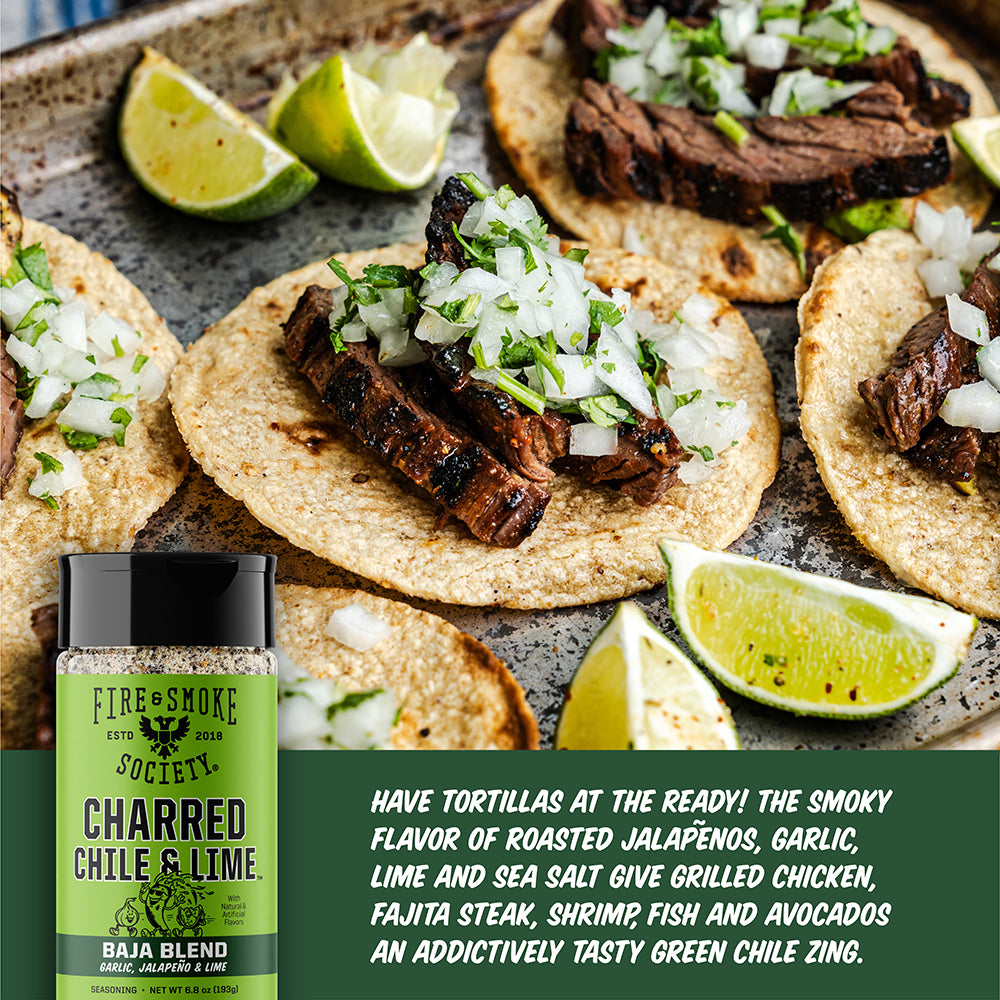 A bottle of Fire & Smoke Society Charred Chile & Lime seasoning is shown in the foreground with grilled steak tacos topped with chopped white onion and lime wedges in the background. A dark green text panel with product copy appears along the bottom right.