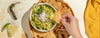 A bowl of guacamole with tortilla chips, garnished with white cheese and cilantro, with a hand dipping a chip into the bowl. The scene includes a lime wedge, a chili pepper, and a yellow cloth on a light surface.