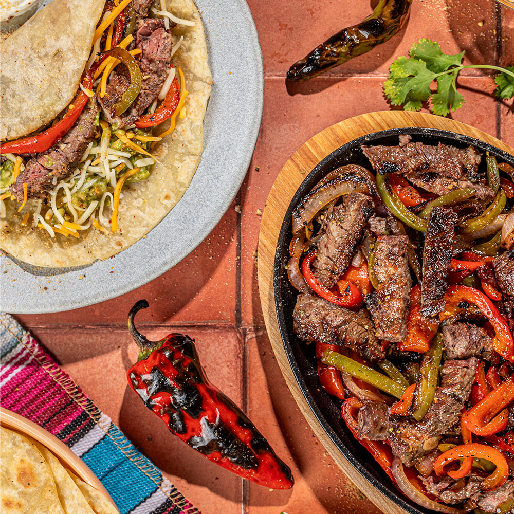Overhead food scene showing a plate of stuffed tortillas and a sizzling skillet of sliced beef fajita with peppers and onions on a terracotta tile surface, with chili peppers, cilantro, and a woven cloth nearby.