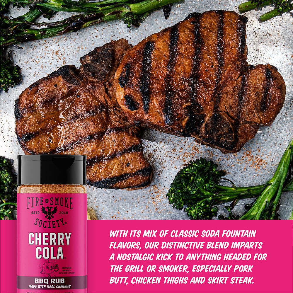 A bottle of Fire & Smoke Cherry Cola BBQ Rub shown next to grilled steak with broccolini. A pink text banner at the bottom describes the flavor and uses.