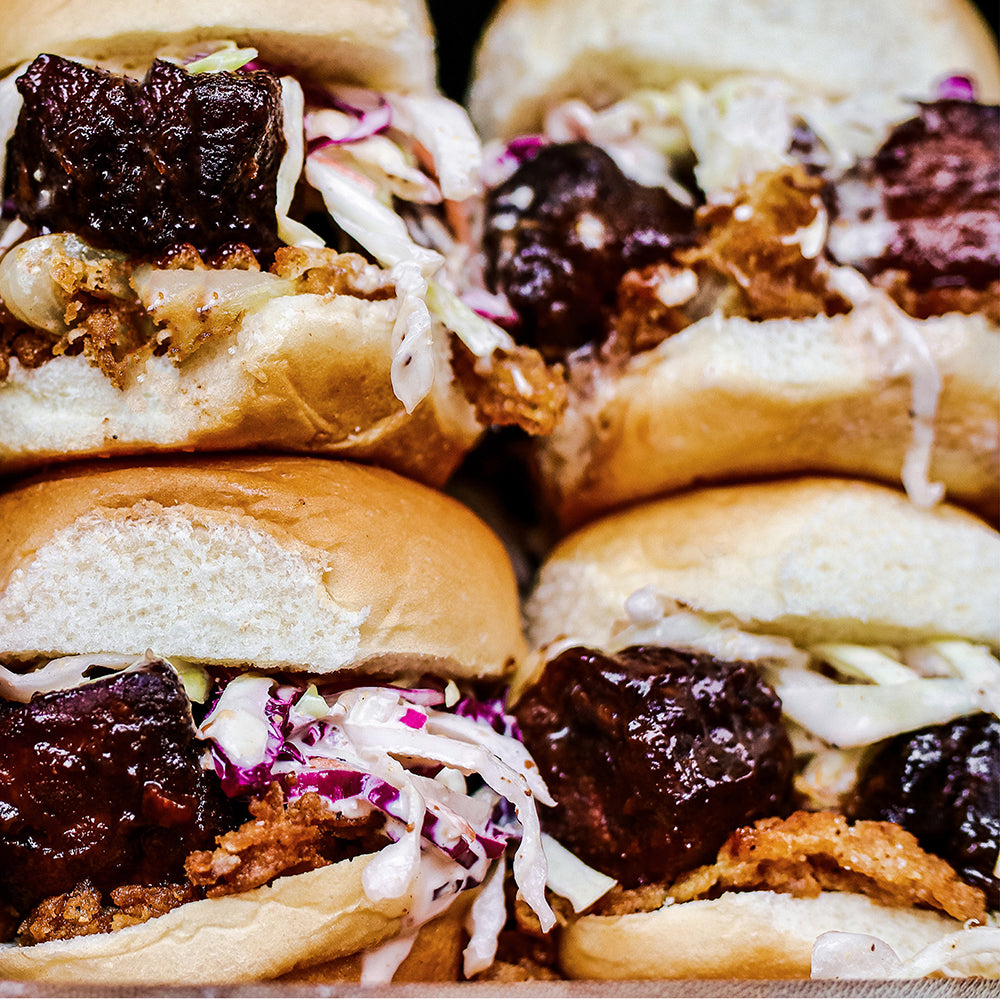 close-up view of four barbecue sliders stacked with crispy fried onions, coleslaw, and glazed meat on buns.