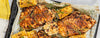 Grilled chicken breasts with corn on the cob, garlic cloves, and herbs arranged on a baking sheet lined with parchment paper.