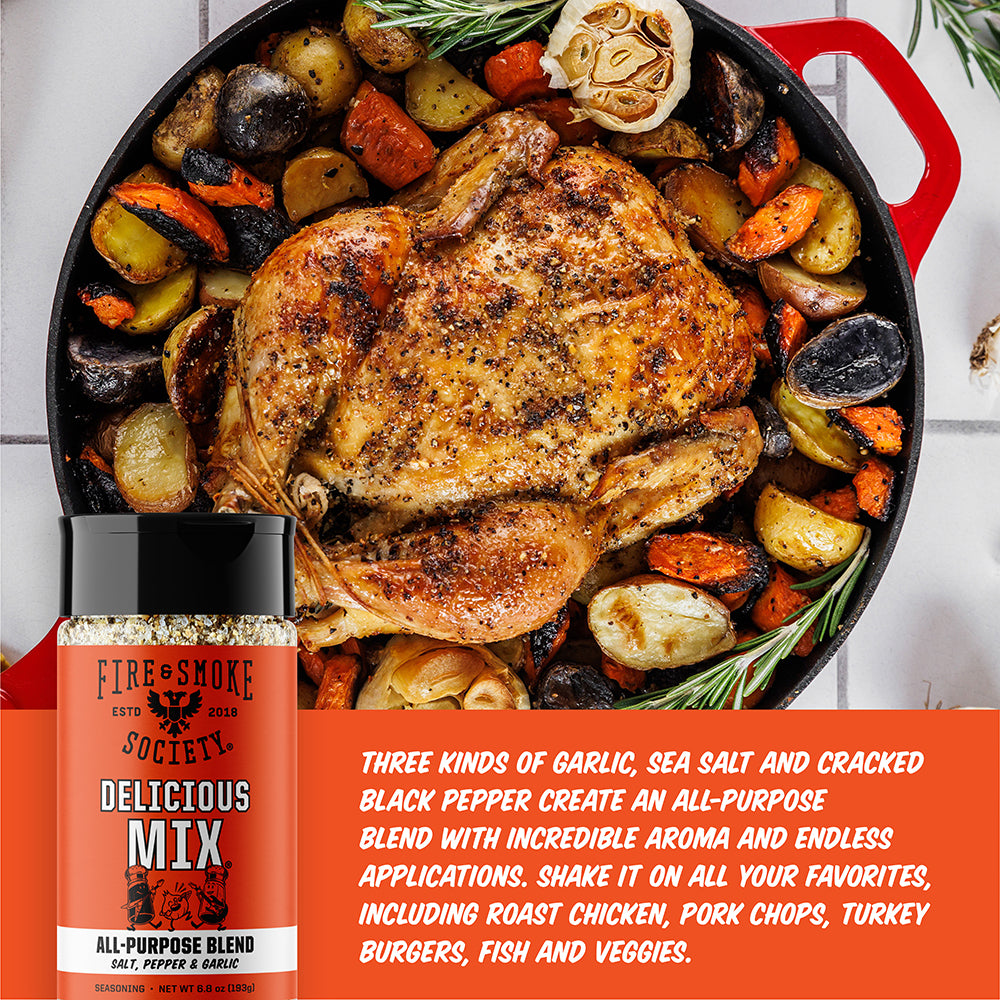 A bottle of Fire & Smoke Society Delicious Mix all-purpose seasoning blend is shown in front of a roasted whole chicken with potatoes, carrots, and garlic in a red cast-iron skillet. White text on an orange banner at the bottom describes the seasoning blend ingredients and uses.