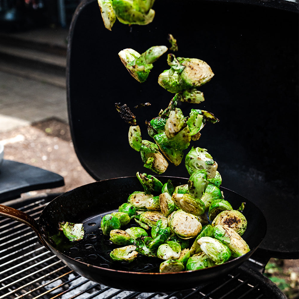 Charred Brussels sprouts falling into a black cast iron skillet on a grill grate