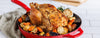 A roasted whole chicken in a red cast-iron skillet with roasted potatoes, carrots, and herbs, set on a beige cloth over a white tiled surface.