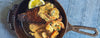 A cast iron skillet filled with a cooked steak, shrimp, creamy sauce, and a lemon wedge, photographed from above on a blue textured fabric background.