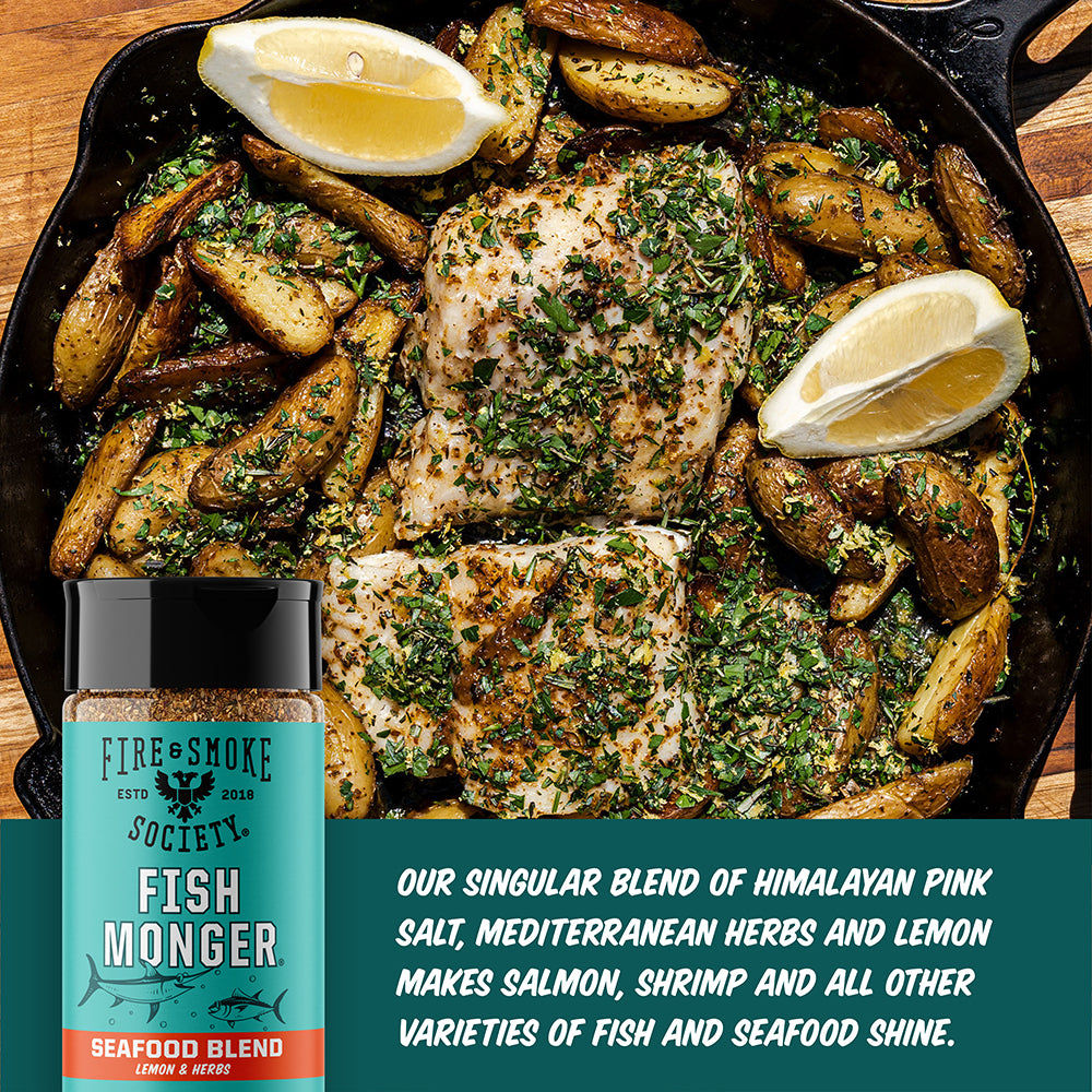 Skillet with seasoned fish and potatoes, lemon wedges, and a bottle of seasoning on a wooden surface.
