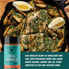 Skillet with seasoned fish and potatoes, lemon wedges, and a bottle of seasoning on a wooden surface.