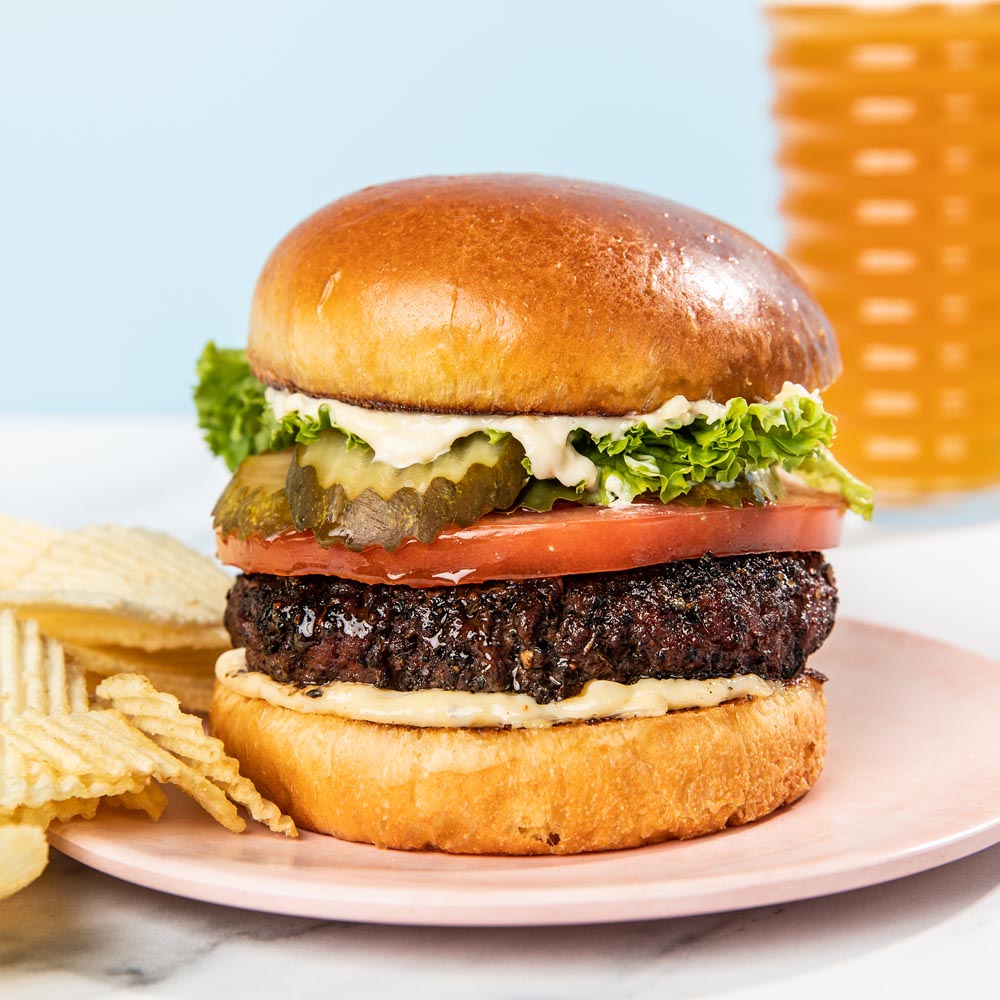 Hamburger with lettuce, tomato, and pickles on a plate with chips and a basket in the background.