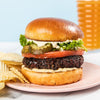 Hamburger with lettuce, tomato, and pickles on a plate with chips and a basket in the background.