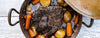 A top-down view of a large cooked pot roast with potatoes and orange carrots simmering in a rustic dark metal pot on a white wooden surface, with part of a metal lid visible at the top right.