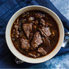 A top-down view of a hearty bowl of beef stew with tender chunks in a rich, dark brown broth, served in a white ceramic bowl on a dark blue textured cloth with a spoon visible on the side.