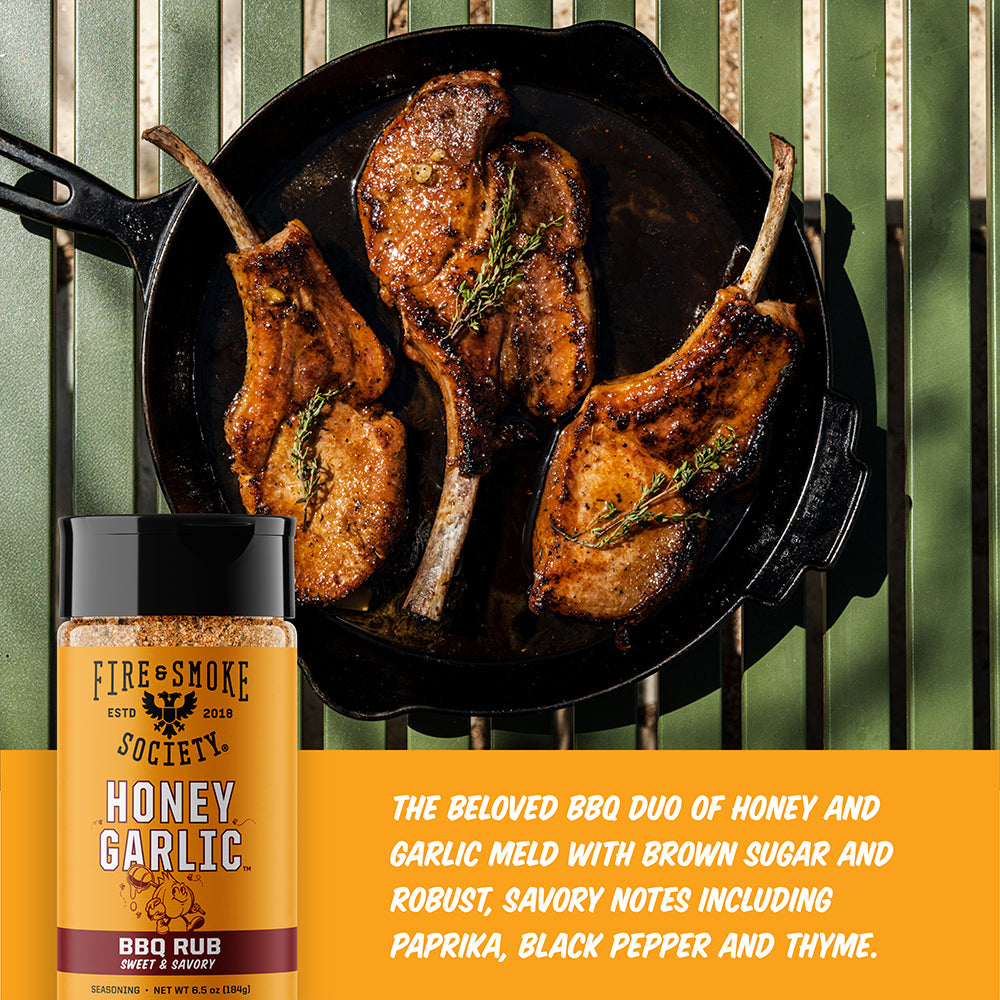 A bottle of Fire and Smoke Society Honey Garlic BBQ Rub is shown next to a black cast iron skillet containing three cooked pork chops on a grill grate, with descriptive text on an orange banner at the bottom.