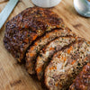 Sliced meatloaf with a glossy browned exterior and visible orange carrot pieces and green herbs in the interior, served on a wooden cutting board with a knife and white plate partially visible.