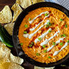 A top-down view of a creamy orange cheese dip in a black cast iron skillet, garnished with ranch dressing drizzle, red hot sauce, and chopped green onions, surrounded by tortilla chips, a jalapeño, and a blue patterned cloth on a dark wooden table.