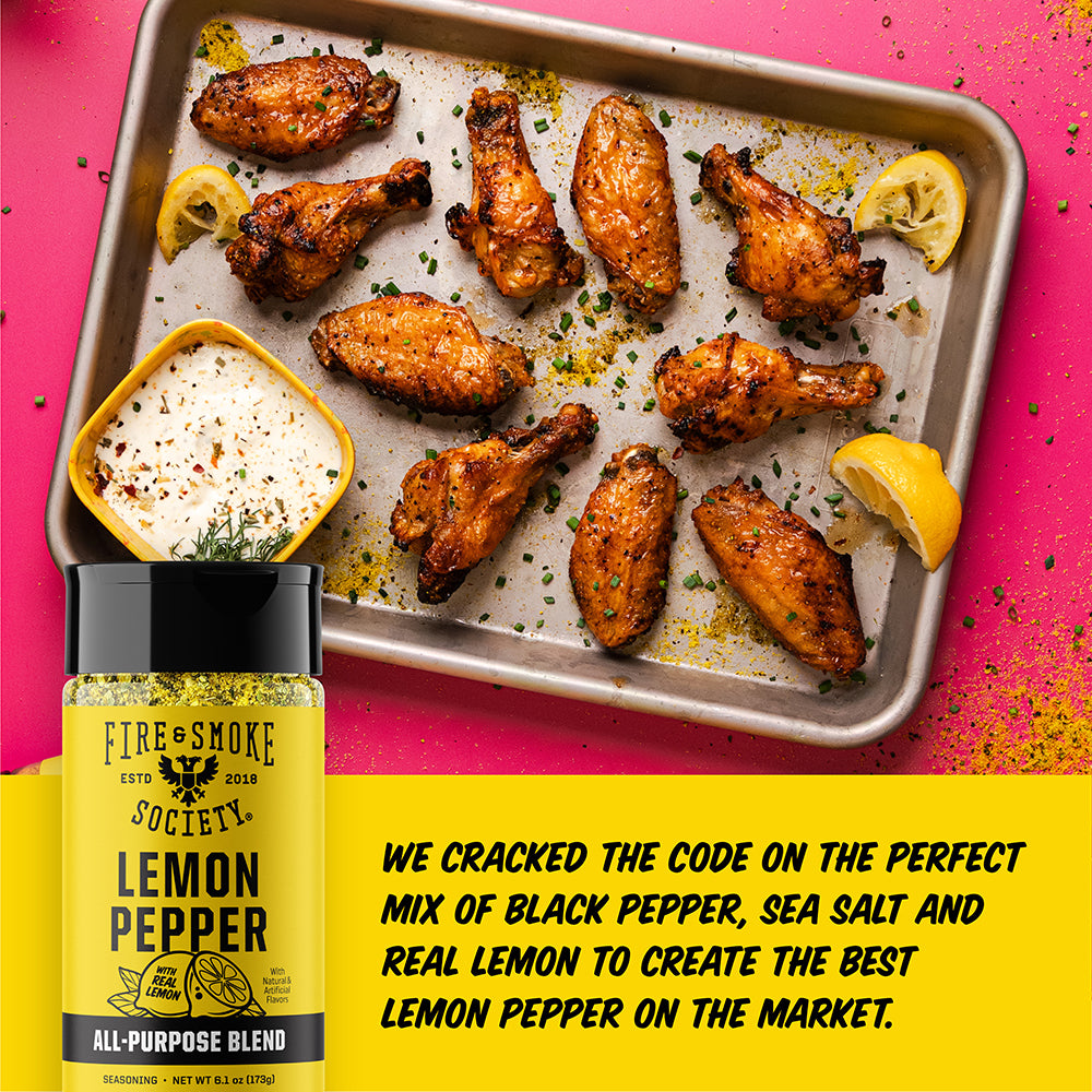 A bottle of Fire & Smoke Society Lemon Pepper seasoning is shown in front of a baking tray of grilled chicken wings with lemon slices and a dipping sauce, on a bright pink background with a yellow text banner.