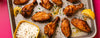 Grilled chicken wings and drumettes on a baking sheet with a side of white dipping sauce, garnished with herbs and lemon wedges, set against a bright pink background.