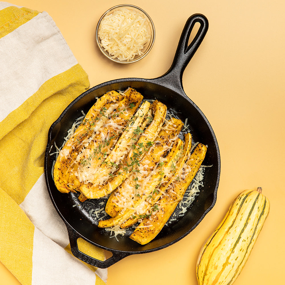 A cast iron skillet filled with baked summer squash strips topped with melted cheese and herbs, arranged on a warm yellow surface with a striped napkin, a small bowl of shredded cheese, and a whole squash nearby.