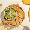 A bowl of guacamole topped with cilantro and crumbled cheese, surrounded by tortilla chips in a peach-colored bowl, photographed from above on a light textured surface with a yellow cloth in the corner.
