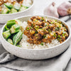 A ceramic bowl filled with white rice topped with étouffée served with green okra pods on the side, with garlic blurred in the background.