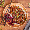 A top-down view of a peach-colored bowl filled with glazed chicken over white rice, garnished with sesame seeds and green onions, served on a wooden table with black chopsticks and a side of red cabbage slaw.