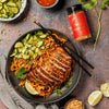 Plated dish of noodles with sliced meat, garnished with vegetables, on a rustic surface with a bottle of Noodle House spice and lime wedges.