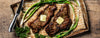Two grilled steaks with melted butter and garlic, served on a rustic baking sheet with green onions, parsley, and a knife on a wooden table.