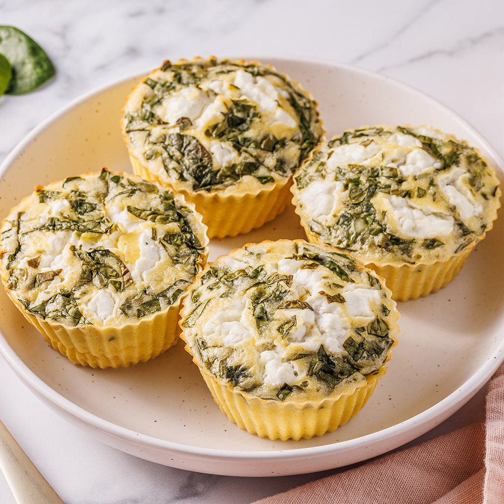 Four baked spinach and cheese egg white muffins in yellow paper cups arranged on a light beige plate, set on a white marble surface with a small green garnish in the corner.