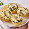 Four baked spinach and cheese egg white muffins in yellow paper cups arranged on a light beige plate, set on a white marble surface with a small green garnish in the corner.