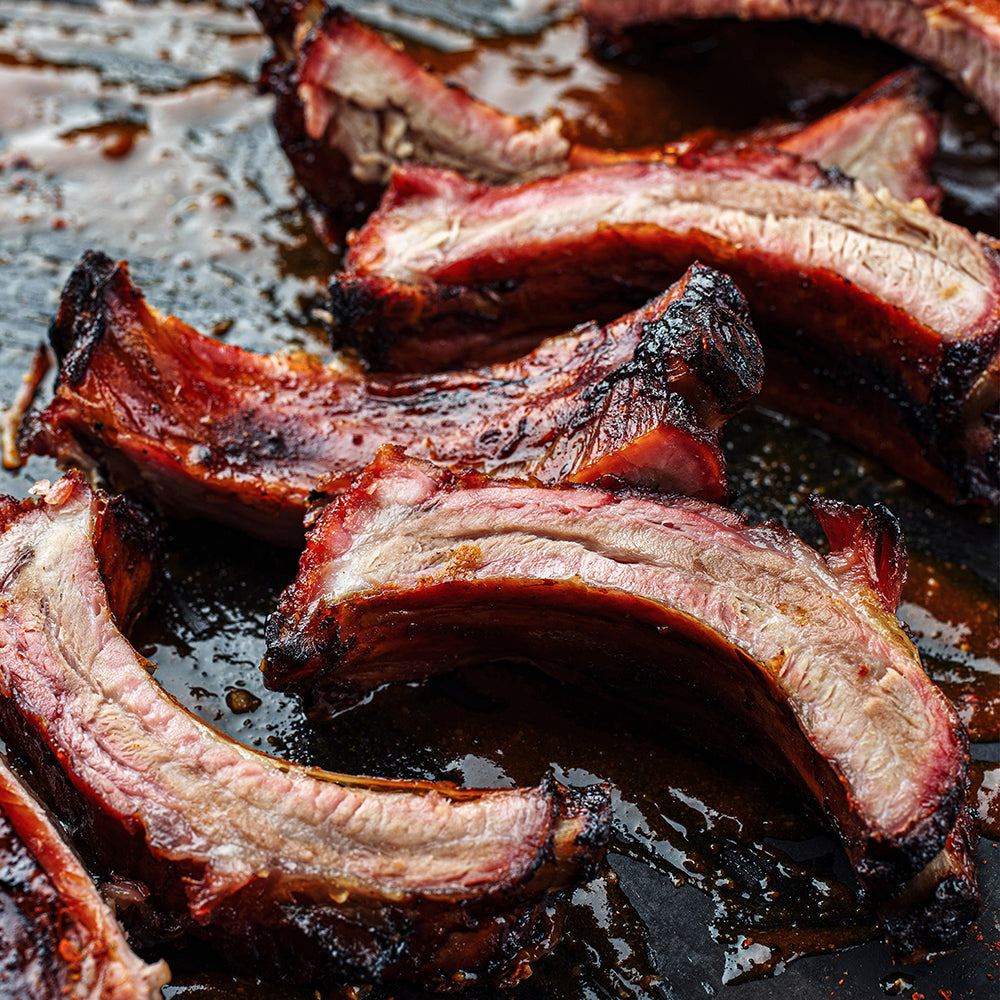 Close-up of barbecued pork ribs with charred edges and a glossy brown sauce, arranged on a dark pan.