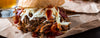 A close-up of a loaded pulled pork sandwich on a bun, topped with shredded coleslaw and dark red barbecue sauce, resting on brown paper wrap with a glass of beer partially visible in the background.