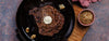 Steak with butter on a black pan on a wooden board, with a bowl of seasoning on a rustic surface.