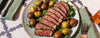 Sliced steak with potatoes and greens on a plate, served with cutlery on a patterned tablecloth.