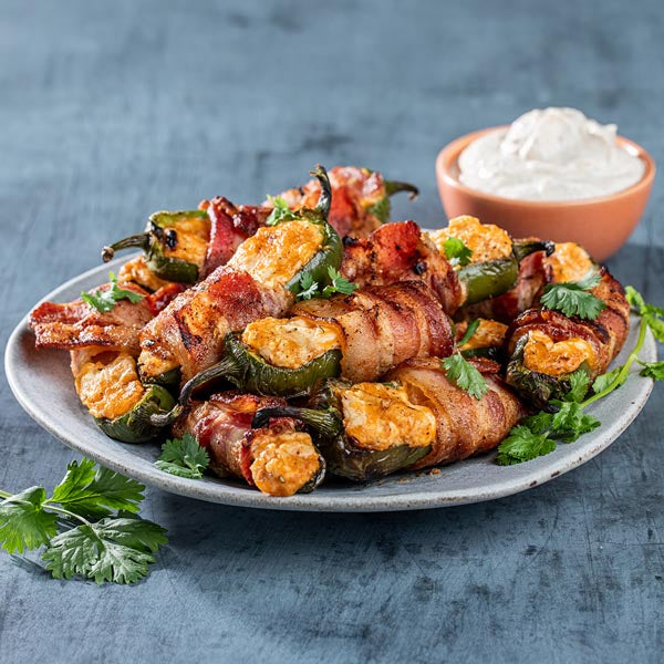 Bacon-wrapped jalapeño poppers on a plate with a side of sour cream.