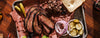 Platter of sliced meat with pickles, onions, and bread on a wooden surface
