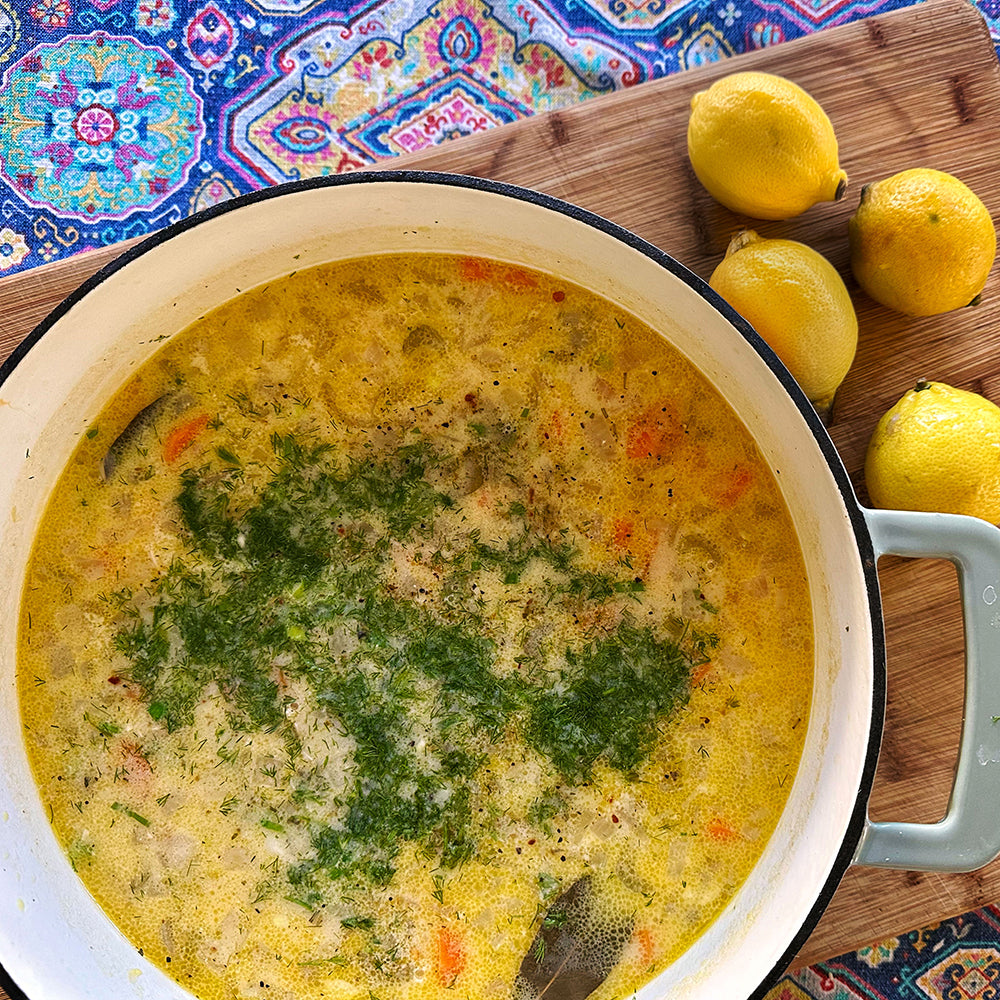 A top-down view of a creamy yellow soup with herbs and carrots in a white enameled cast iron pot, placed on a wooden cutting board with lemons and a colorful patterned cloth underneath.