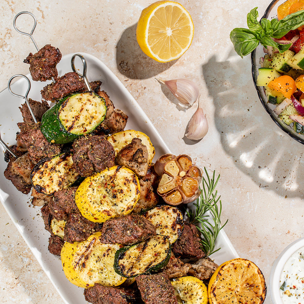 Grilled meat and vegetable skewers on a white serving platter, arranged on a light stone surface with a lemon half, garlic cloves, a bowl of salad, and a small dipping dish nearby.