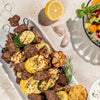 Grilled meat and vegetable skewers on a white serving platter, arranged on a light stone surface with a lemon half, garlic cloves, a bowl of salad, and a small dipping dish nearby.