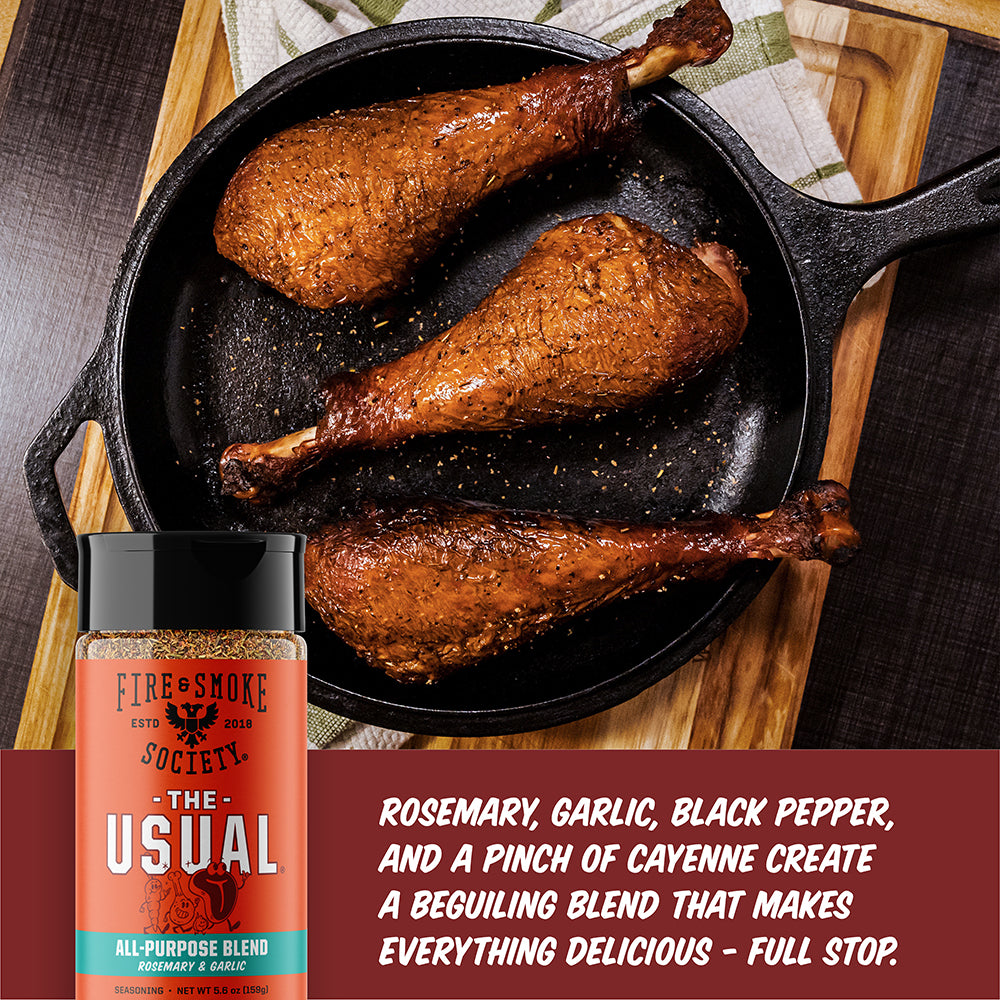 A bottle of Fire Smoke Society 'The Usual' all-purpose spice blend is shown in the foreground next to three cooked chicken drumsticks in a black cast-iron skillet. The background features a wooden cutting board and a dark textured surface, with a red text banner at the bottom describing the seasoning blend.