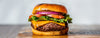 A close-up of a gourmet cheeseburger on a wooden board, featuring a juicy beef patty, melted cheddar cheese, fresh green lettuce, red onion, tomato, and a toasted bun.
