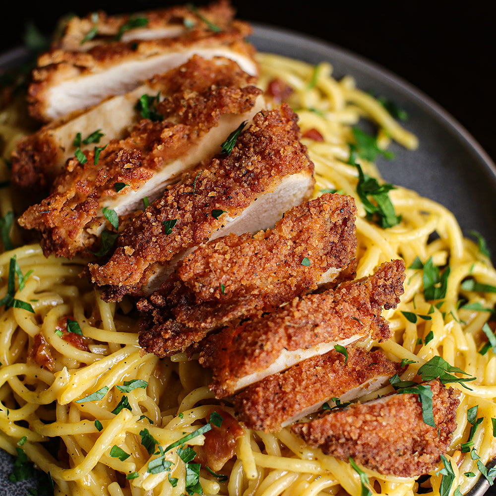 Sliced crispy breaded chicken served over creamy pasta with chopped parsley garnish, presented on a dark plate against a dark background.