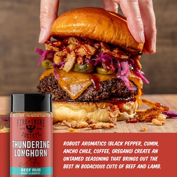 Huge burger with various toppings and a bottle of 'Thundering Longhorn' beef rub on a wooden surface.